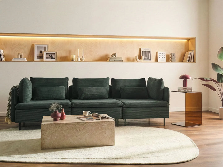 A modern dark green sectional sofa in a styled living room setting, featuring a matching dark green throw pillow, a light wood coffee table, a cream rug, and a built-in wooden shelf with warm lighting on the wall behind.