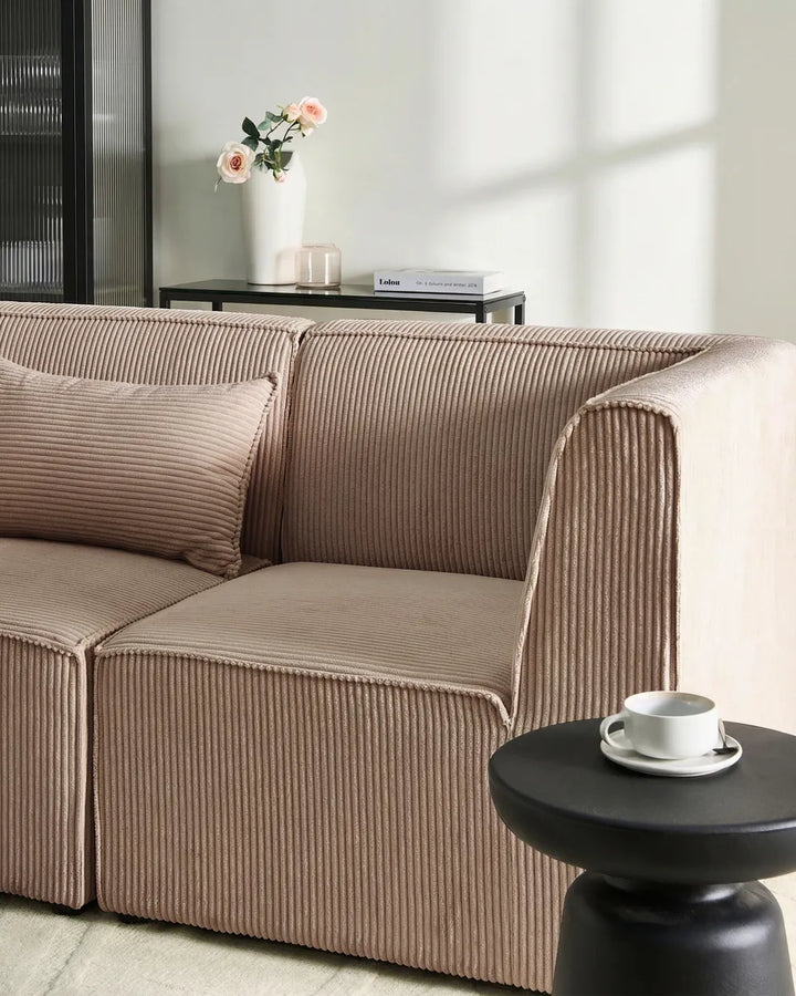 A modern corduroy sofa in a warm taupe-brown color, styled in a minimalist living room with a matching throw pillow, a small black side table with a white cup, and a side table in the background holding a vase of pink flowers.