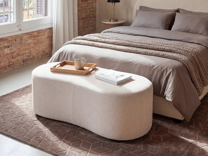 A modern curved upholstered ottoman bench in a warm cream or light beige fabric, styled in a contemporary bedroom setting with a bed, rug, and window in the background.