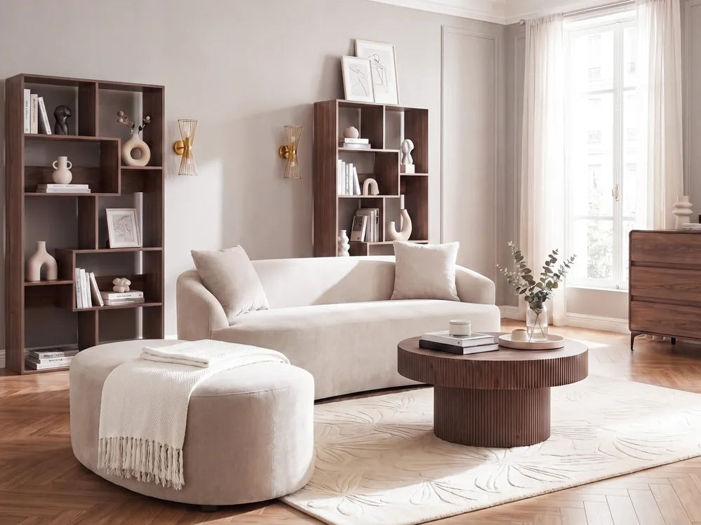 A modern living room featuring a light beige fabric sofa, a matching round ottoman with a white throw blanket, a round dark wood coffee table, and dark wood shelving units against neutral walls.