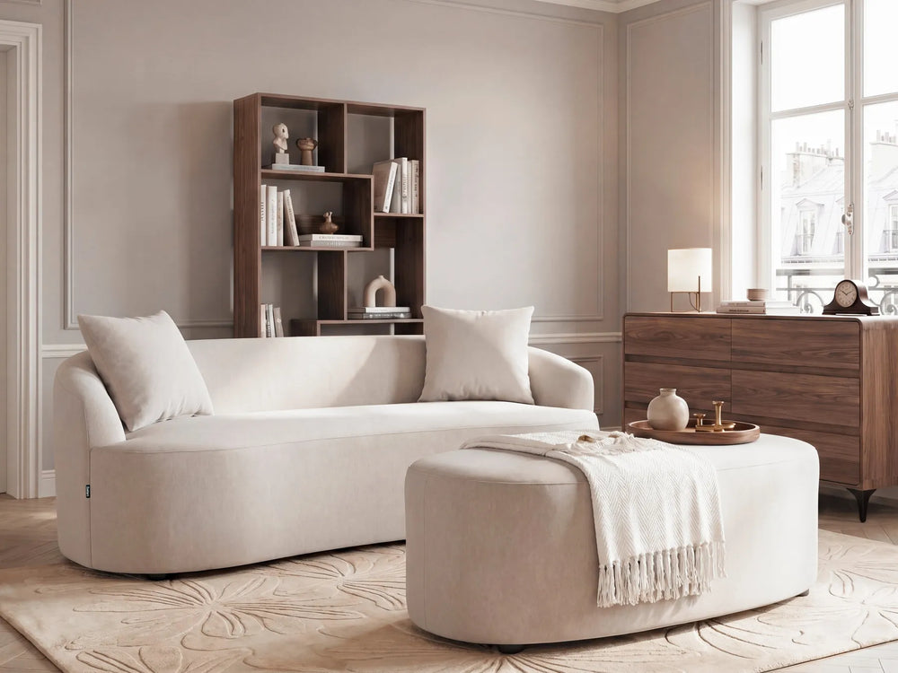 A modern beige sectional sofa with matching throw pillows in a styled living room, paired with a matching round ottoman and a dark wood shelving unit in the background.