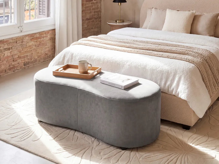 A gray upholstered oval ottoman or bench placed at the foot of a bed in a softly lit bedroom. The room includes cream bedding, a patterned rug, exposed brick wall, window, and bedside lamp.