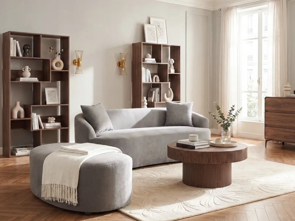 A modern living room setup featuring a light gray fabric sofa, a matching round ottoman with a white throw blanket, a round wooden coffee table, and dark wood shelving units against a cream-colored wall.