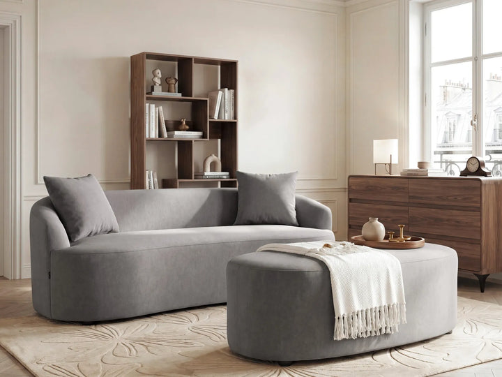 A modern gray fabric sofa with matching throw pillows and a matching round ottoman in a bright, elegant living room with wood furniture and a window.