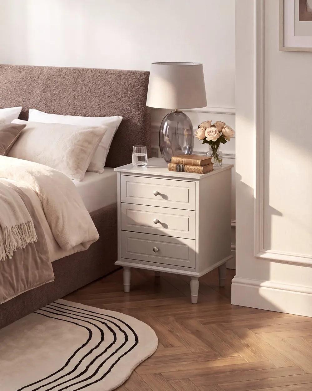 A light gray three-drawer nightstand in a styled bedroom setting, styled with a table lamp, books, and a vase of flowers next to a bed with a brown upholstered headboard.
