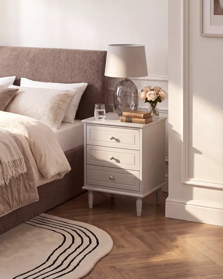 A light gray three-drawer nightstand in a styled bedroom setting, styled with a table lamp, books, and a vase of flowers next to a bed with a brown upholstered headboard.