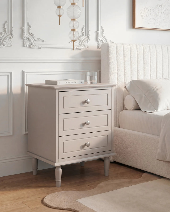A light beige three-drawer nightstand with silver knobs stands beside a bed in a bright, neutral bedroom setting.