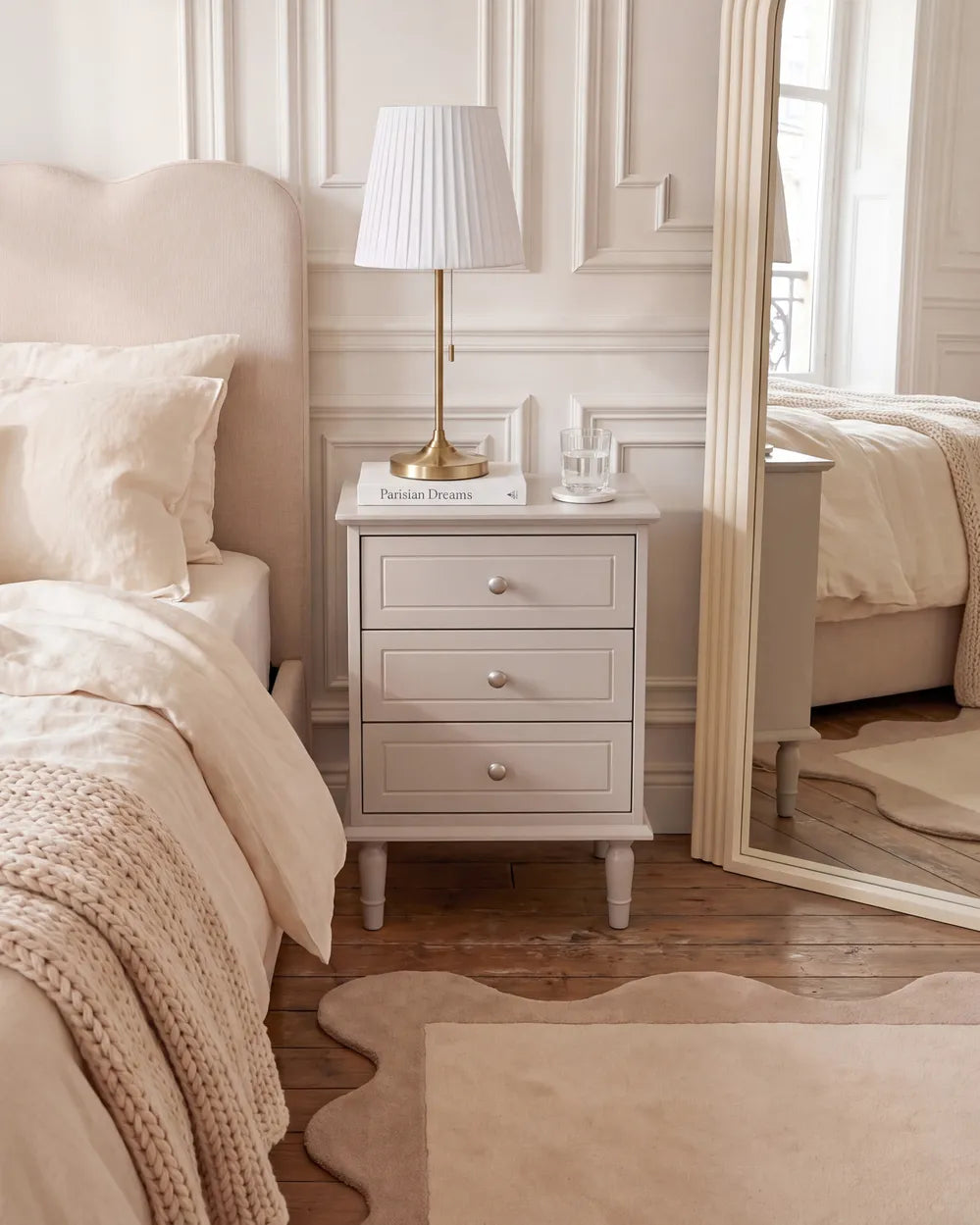 A light gray three-drawer nightstand in a beautifully styled bedroom, featuring a white pleated table lamp with a gold base, a small book, and a glass cup on top. The nightstand is positioned next to a bed with beige bedding and a large floor mirror reflecting the room.