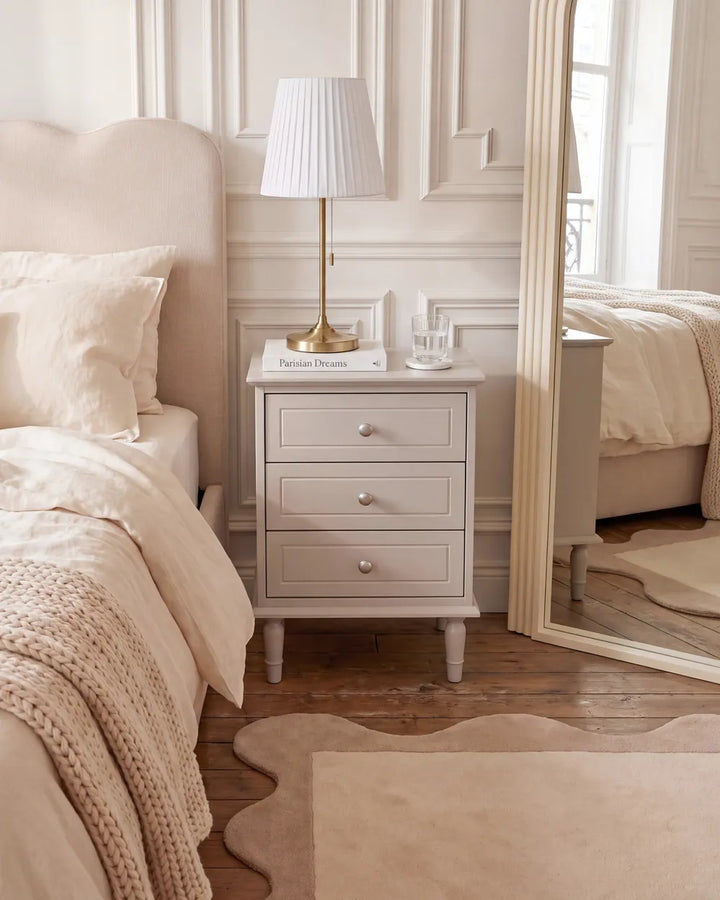 A light gray three-drawer nightstand in a beautifully styled bedroom, featuring a white pleated table lamp with a gold base, a small book, and a glass cup on top. The nightstand is positioned next to a bed with beige bedding and a large floor mirror reflecting the room.