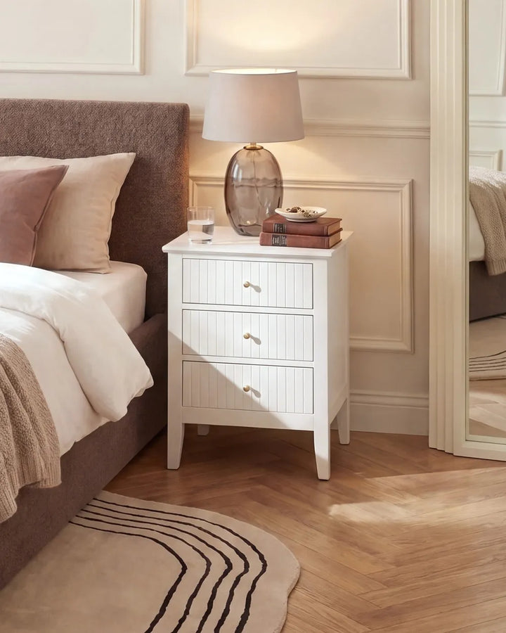 A white three-drawer nightstand with a shiplap-style front and small gold knobs, styled in a bedroom next to a bed with a brown upholstered headboard. A glass table lamp and stacked books sit on top of the nightstand, which is set on a light wood floor.