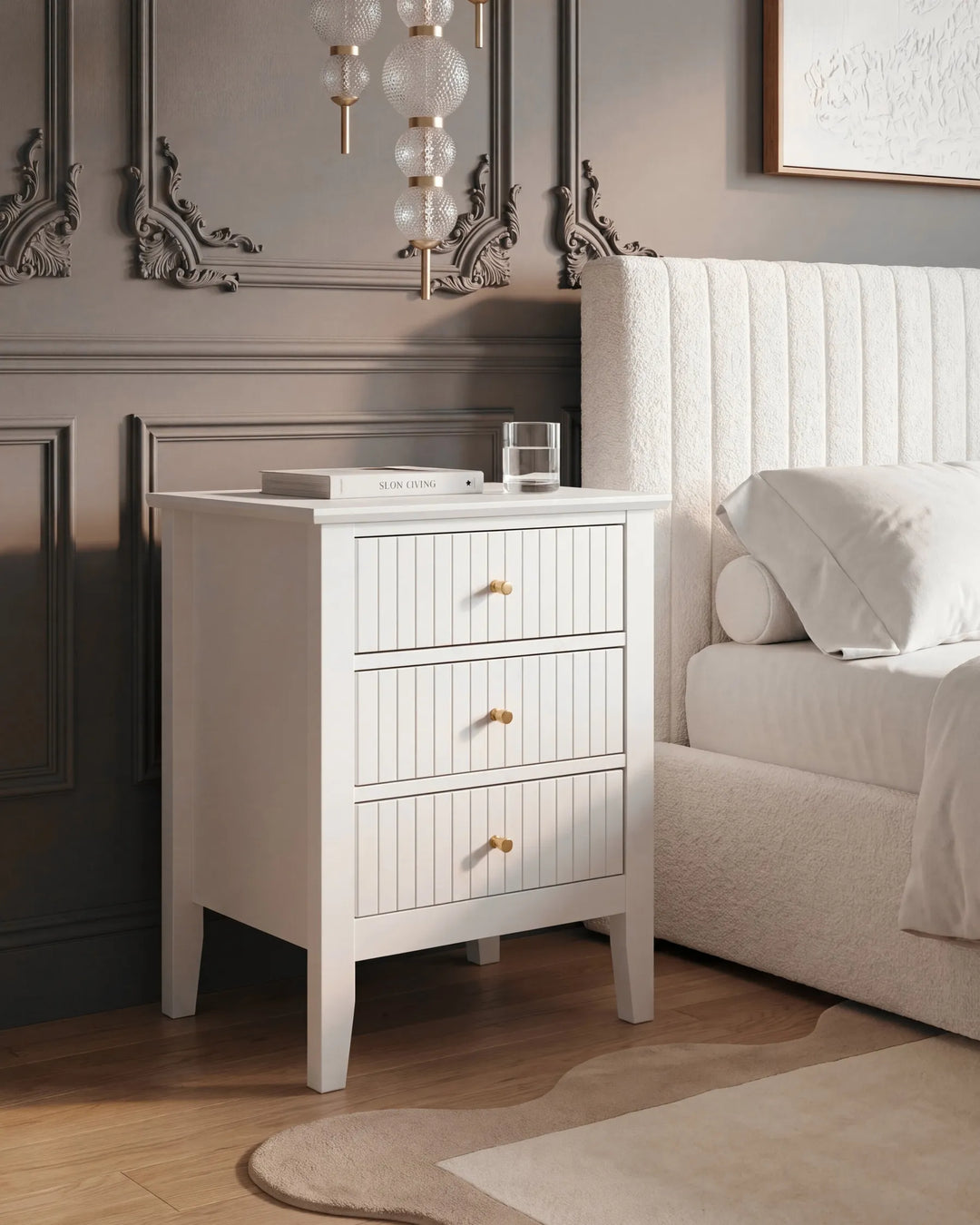 A white three-drawer nightstand with gold knobs stands beside a bed in a styled bedroom. The nightstand has a vertical paneled front, tapered legs, and is shown with a glass and a book on top.