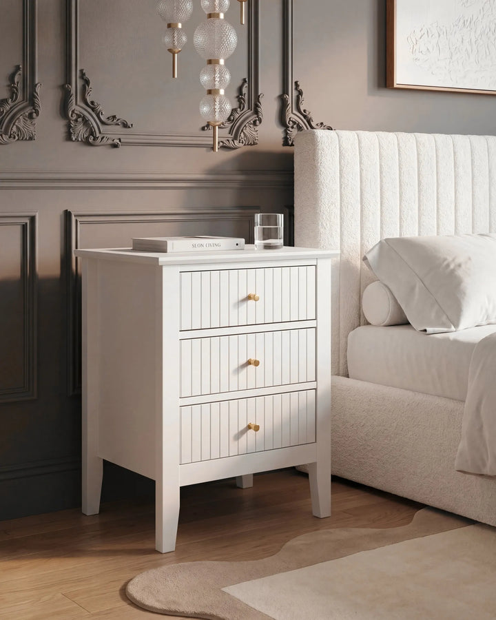 A white three-drawer nightstand with gold knobs stands beside a bed in a styled bedroom. The nightstand has a vertical paneled front, tapered legs, and is shown with a glass and a book on top.