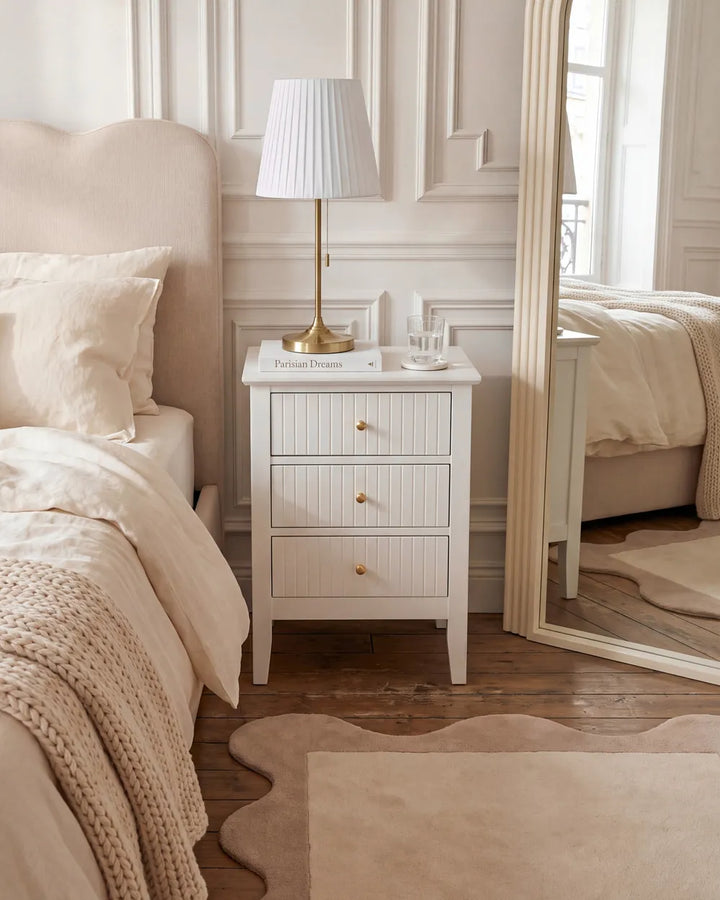 A white three-drawer nightstand with gold knobs in a beautifully styled bedroom, featuring a white pleated lamp, a glass of water, and a white 'Paisley Designs' logo plaque on top.