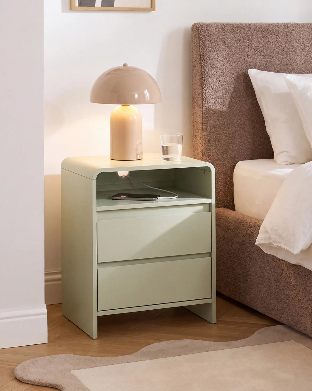 A light sage green three-drawer nightstand in a bedroom setting, styled with a beige dome lamp and a glass of water on top, next to a bed with a brown upholstered headboard.