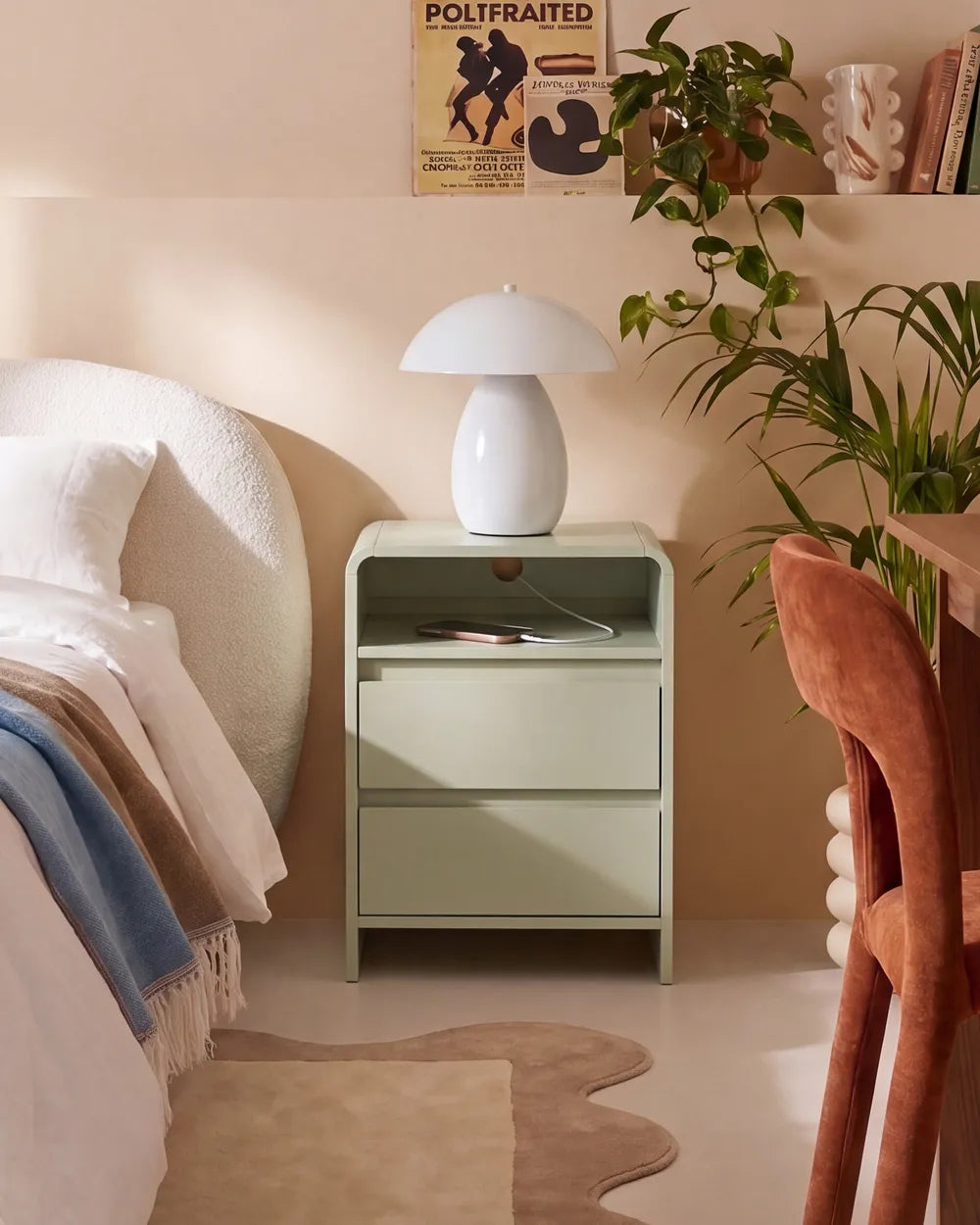 A light sage green nightstand with two drawers in a cozy bedroom setting, topped with a white mushroom-shaped table lamp. The scene includes a bed with white and blue bedding, a brown velvet chair, a potted green plant, and a beige rug on the floor.