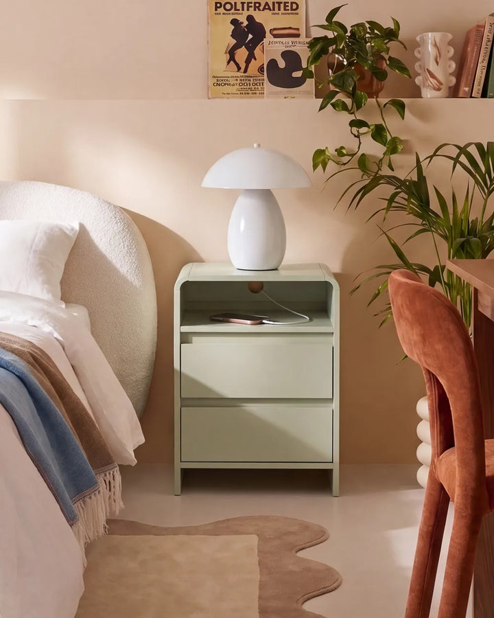 A light sage green nightstand with two drawers in a cozy bedroom setting, topped with a white mushroom-shaped table lamp. The scene includes a bed with white and blue bedding, a brown velvet chair, a potted green plant, and a beige rug on the floor.