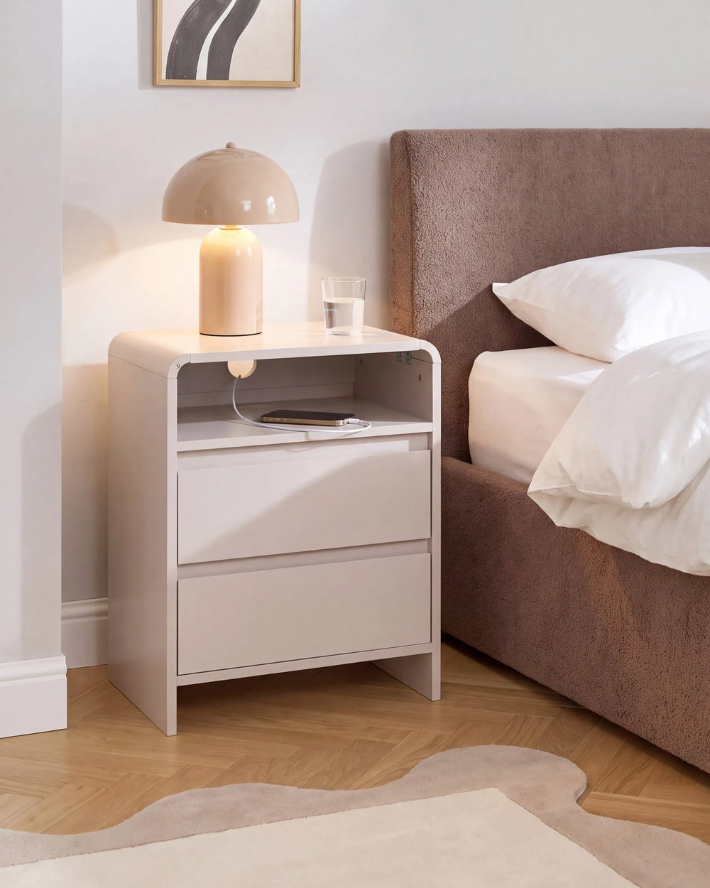 A modern white nightstand with two drawers and an open shelf, styled in a bedroom next to a bed with a brown headboard. A beige dome lamp and a glass of water sit on top.