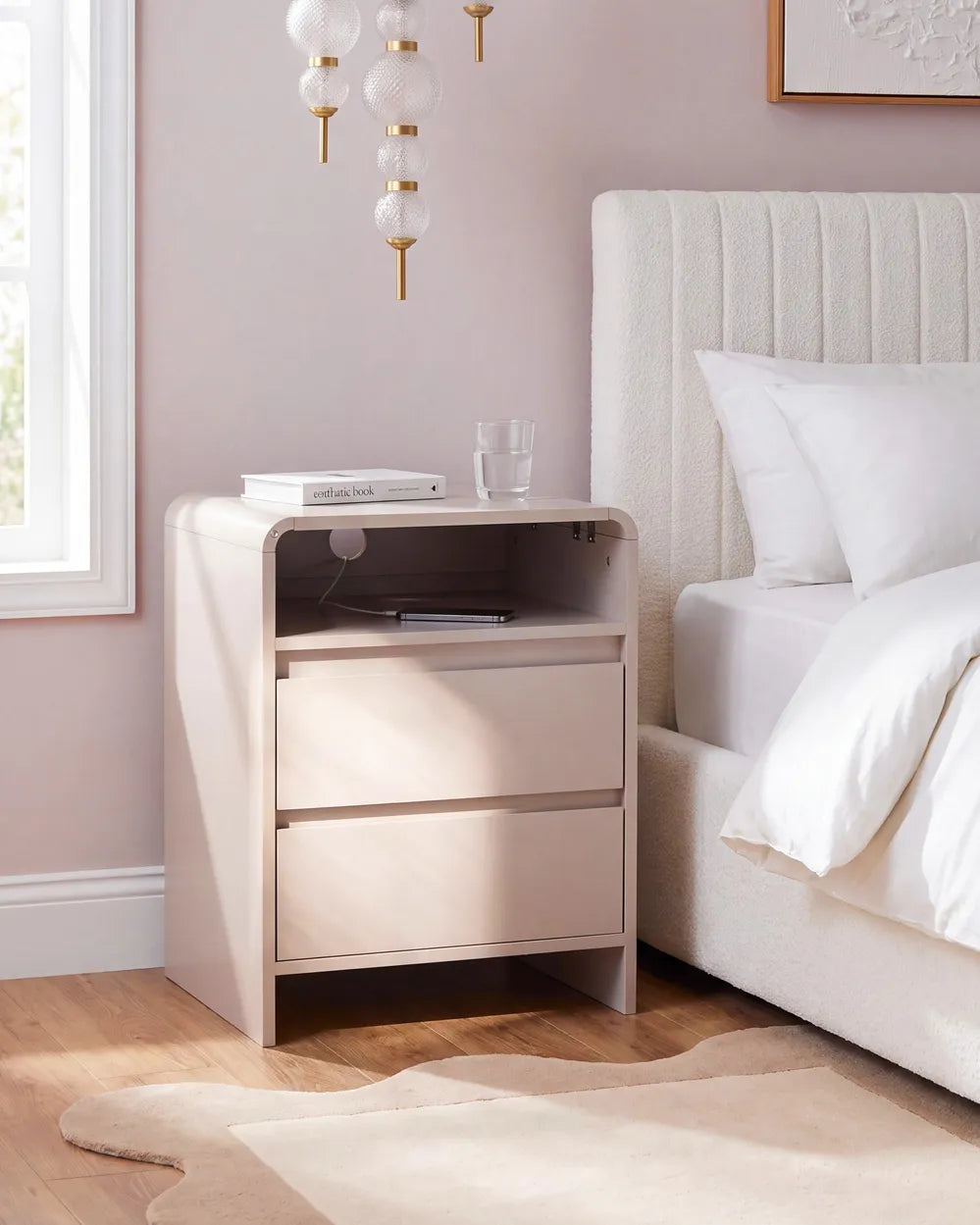 A modern, minimalist two-drawer nightstand in a light blush pink finish, styled next to a bed with white linens in a bedroom setting.