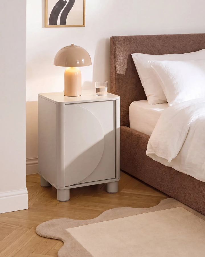 A modern light gray nightstand with a curved front detail and cylindrical legs, styled in a bedroom next to a bed with white linens and a brown upholstered headboard. A beige dome lamp and a small glass of water sit on top.