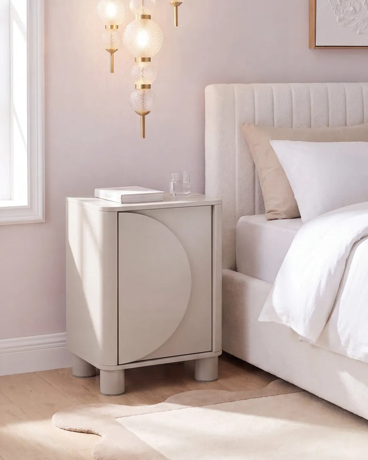 A modern cream-colored nightstand with a curved front detail, positioned next to a bed in a softly lit bedroom with a tufted headboard, white bedding, and a hanging glass globe lamp.