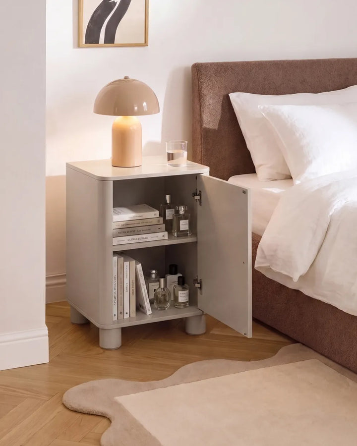 A light gray bedside cabinet with an open door, styled next to a bed with white linens and a brown upholstered headboard. A beige dome lamp sits on top of the cabinet, which also holds books and small bottles.