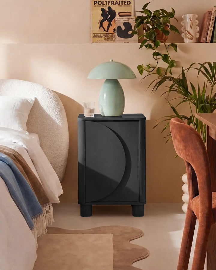 A modern black nightstand with a curved crescent moon design on the front, styled in a warm beige bedroom next to a bed, holding a light green mushroom lamp.