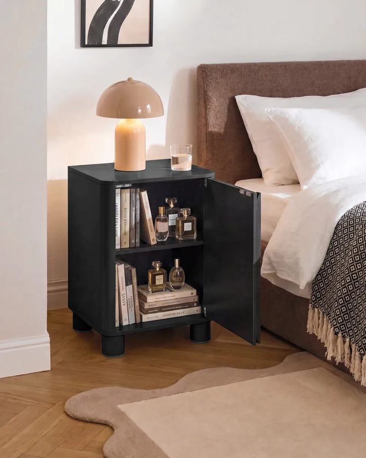 A black nightstand with an open door, styled in a bedroom next to a bed with white linens. The nightstand holds books, perfume bottles, a lit dome lamp, and a small glass of water.