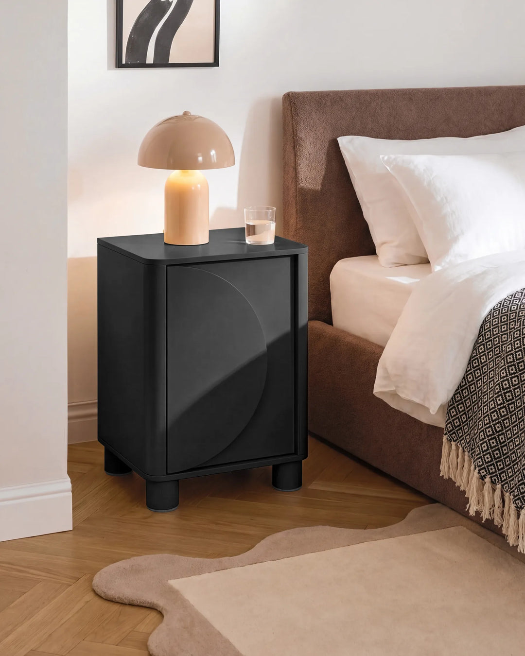 A modern black nightstand with a curved front detail, styled in a bedroom next to a bed with white linens and a brown upholstered headboard. A beige dome lamp and a small glass of water sit on top.