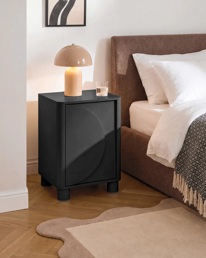 A modern black nightstand with a curved front detail, styled in a bedroom next to a bed with white linens and a brown upholstered headboard. A beige dome lamp and a small glass of water sit on top.