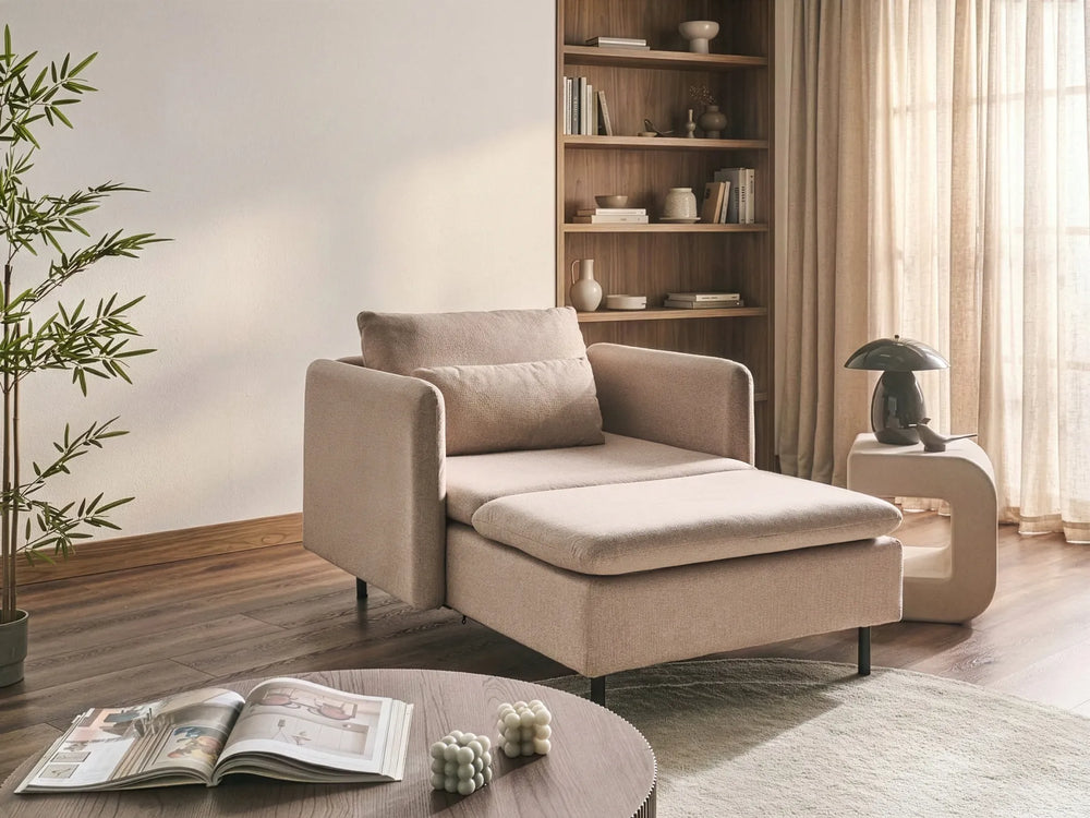 A modern beige chaise lounge chair with a matching cushion, styled in a warm, minimalist living room with a wooden bookshelf, curtains, and a round coffee table in the foreground.