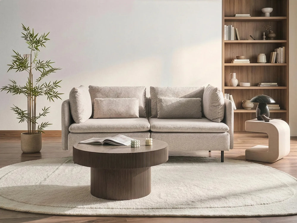 A modern living room scene featuring a light beige upholstered sofa with matching pillows, a round dark wood coffee table, a large textured light rug, a potted bamboo plant, and a tall wooden bookshelf in the background.