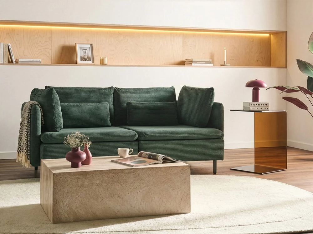 A modern living room setting featuring a dark green textured fabric sofa as the main product, styled with a beige stone coffee table, a white rug, and warm wood shelving in the background.