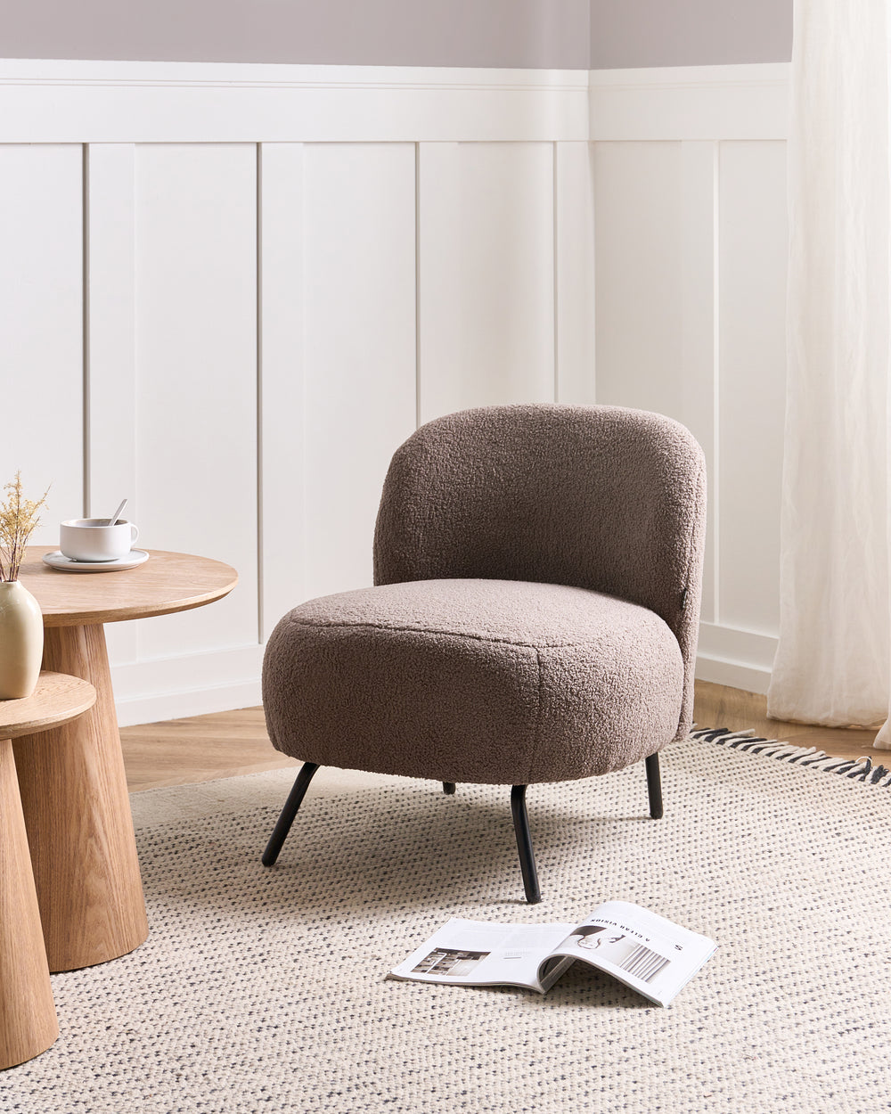 A round upholstered accent chair in a textured taupe bouclé fabric with black tapered legs, set in a modern living room corner with a wooden side table, woven rug, and magazine on the floor.