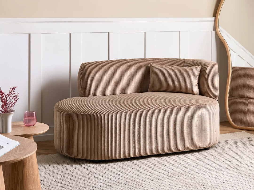A modern curved lounge chair upholstered in beige corduroy fabric, styled in a minimalist living room setting with a white paneled wall, a small wooden side table, and a textured rug.