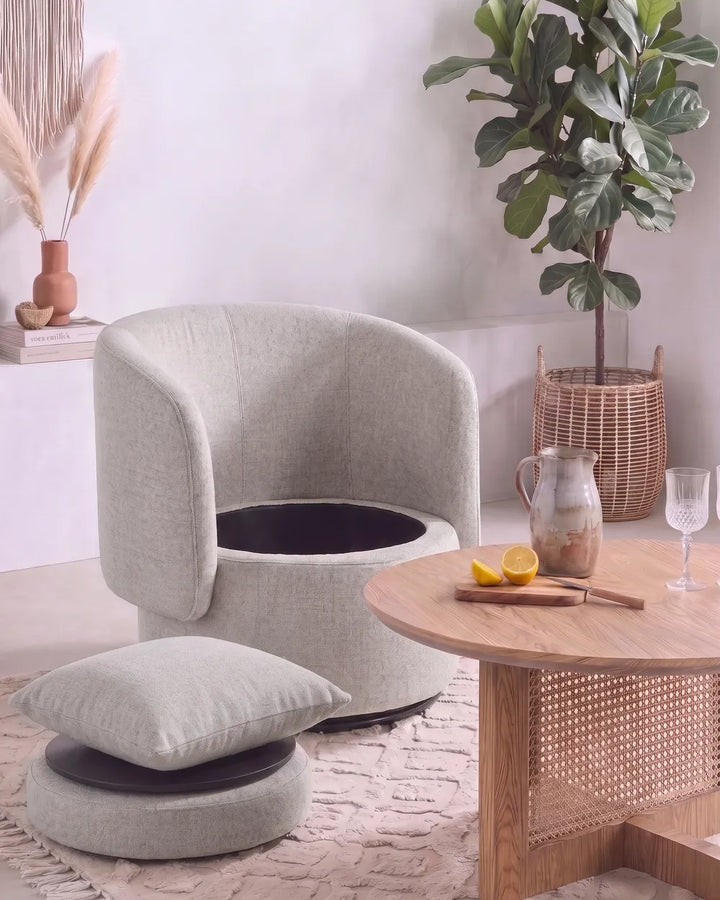 A light beige upholstered barrel-style chair with a matching cushioned ottoman in a styled room setting next to a wooden table with a pitcher and lemon slices.