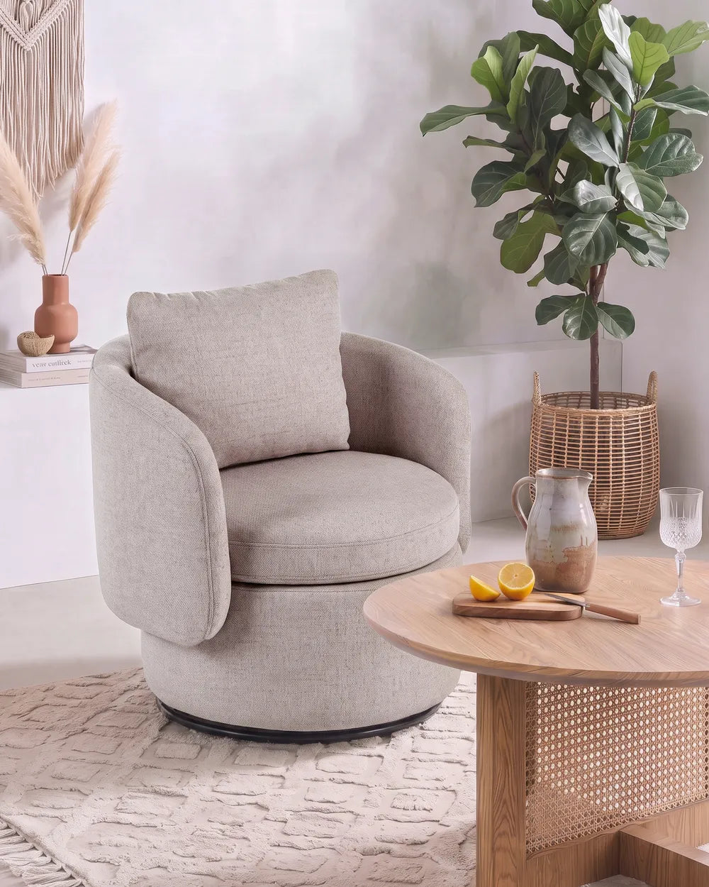 A round upholstered accent chair in a light beige fabric with a matching back cushion, styled in a bright, modern room next to a wooden coffee table and a potted fiddle-leaf fig plant.