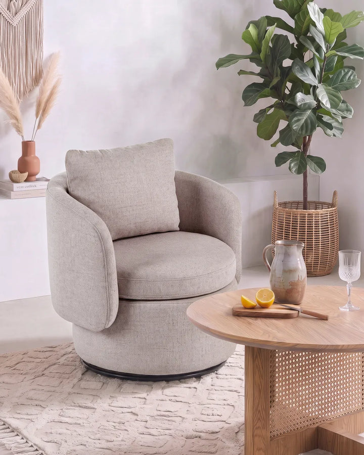 A round upholstered accent chair in a light beige fabric with a matching back cushion, styled in a bright, modern room next to a wooden coffee table and a potted fiddle-leaf fig plant.
