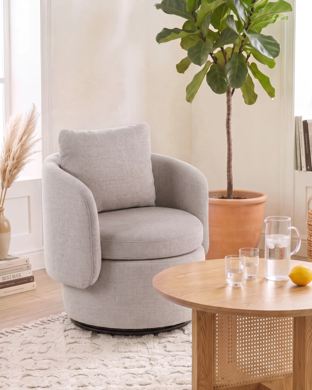 A light gray upholstered swivel armchair with a matching back cushion, shown in a bright living room setting next to a wooden coffee table and a potted fiddle-leaf fig plant.