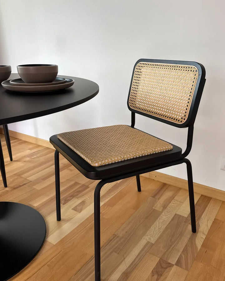 A modern dining chair with a black metal frame and natural woven cane backrest and seat, shown in a minimalist room setting with a wooden floor and a black table.
