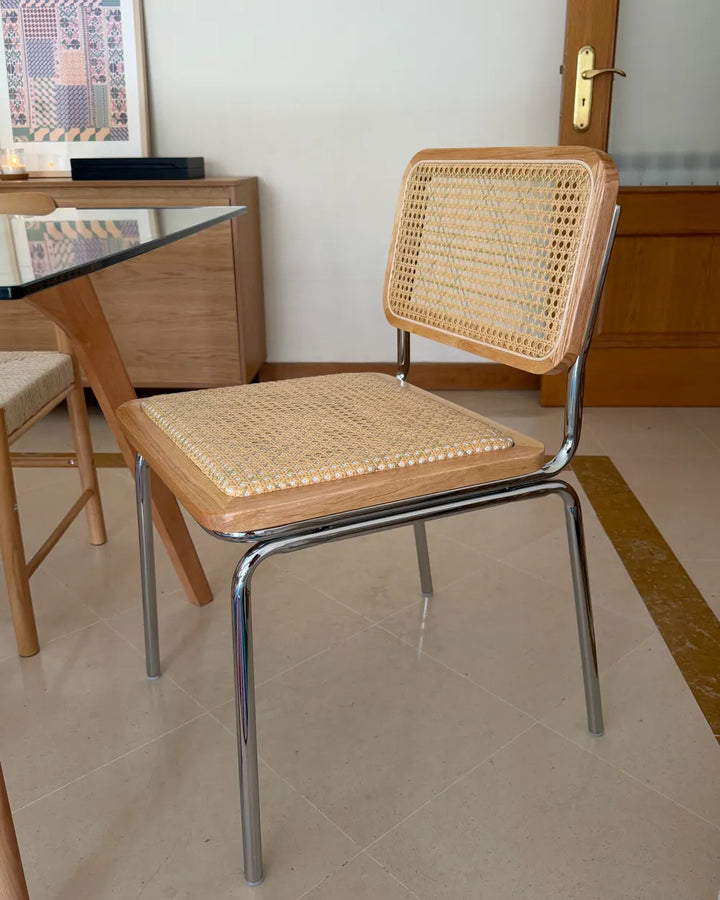 A mid-century modern dining chair with a light wood frame, woven cane backrest, and seat cushion, supported by a chrome tubular steel frame, shown in a dining room setting.