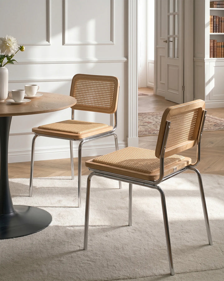 Two modern dining chairs with woven cane seats and backs and chrome metal legs, shown in a bright, sunlit dining room next to a round wooden table.
