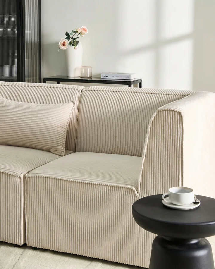 A modern beige corduroy sofa with matching pillows in a bright, minimalist living room setting, featuring a small black side table with a white cup in the foreground.