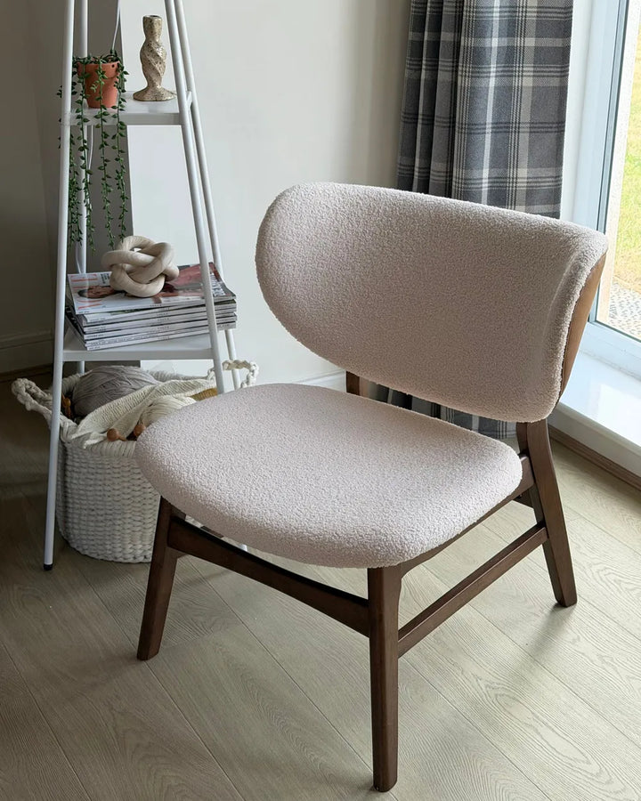A modern accent chair with a textured cream bouclé seat and back cushion on a dark wood frame, positioned in a bright room next to a window with plaid curtains.
