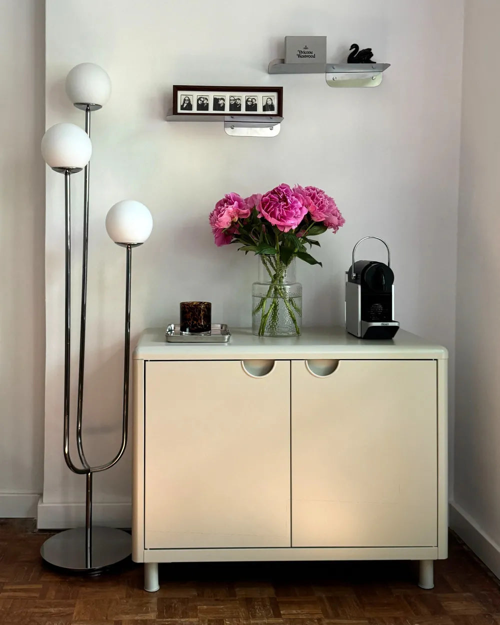 A cream-colored two-door cabinet styled in a minimalist room corner, featuring a vase of pink flowers, a black coffee machine, and a tall chrome floor lamp with white globe shades.
