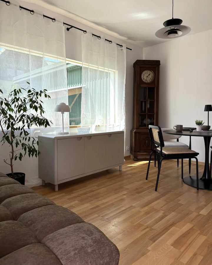 A modern living room interior featuring light oak wood floors, white sheer curtains, a gray sideboard cabinet, a dark wood grandfather clock, and a dining area with a black table and chairs.