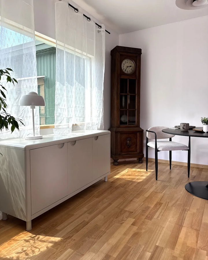 A modern interior room with light wood floors, white walls, sheer white curtains, a white sideboard cabinet, a dark wood grandfather clock, a small dining table with a white chair, and a potted plant.