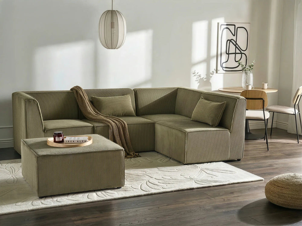 An L-shaped sectional sofa in a muted sage green color, styled in a modern living room with a matching ottoman, a beige throw blanket, a patterned rug, and dark wood floors.