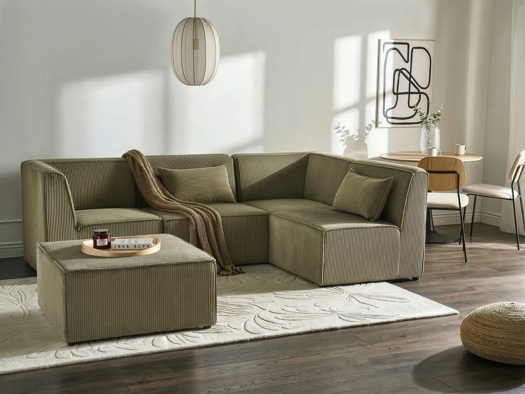 An L-shaped sectional sofa in a muted sage green color, styled in a modern living room with a matching ottoman, a beige throw blanket, a patterned rug, and dark wood floors.