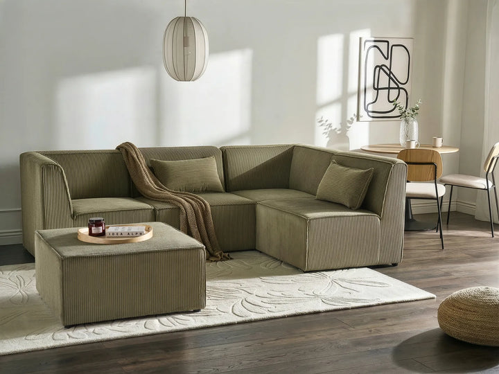 An L-shaped sectional sofa in a muted sage green color, styled in a modern living room with a matching ottoman, a beige throw blanket, a patterned rug, and dark wood floors.