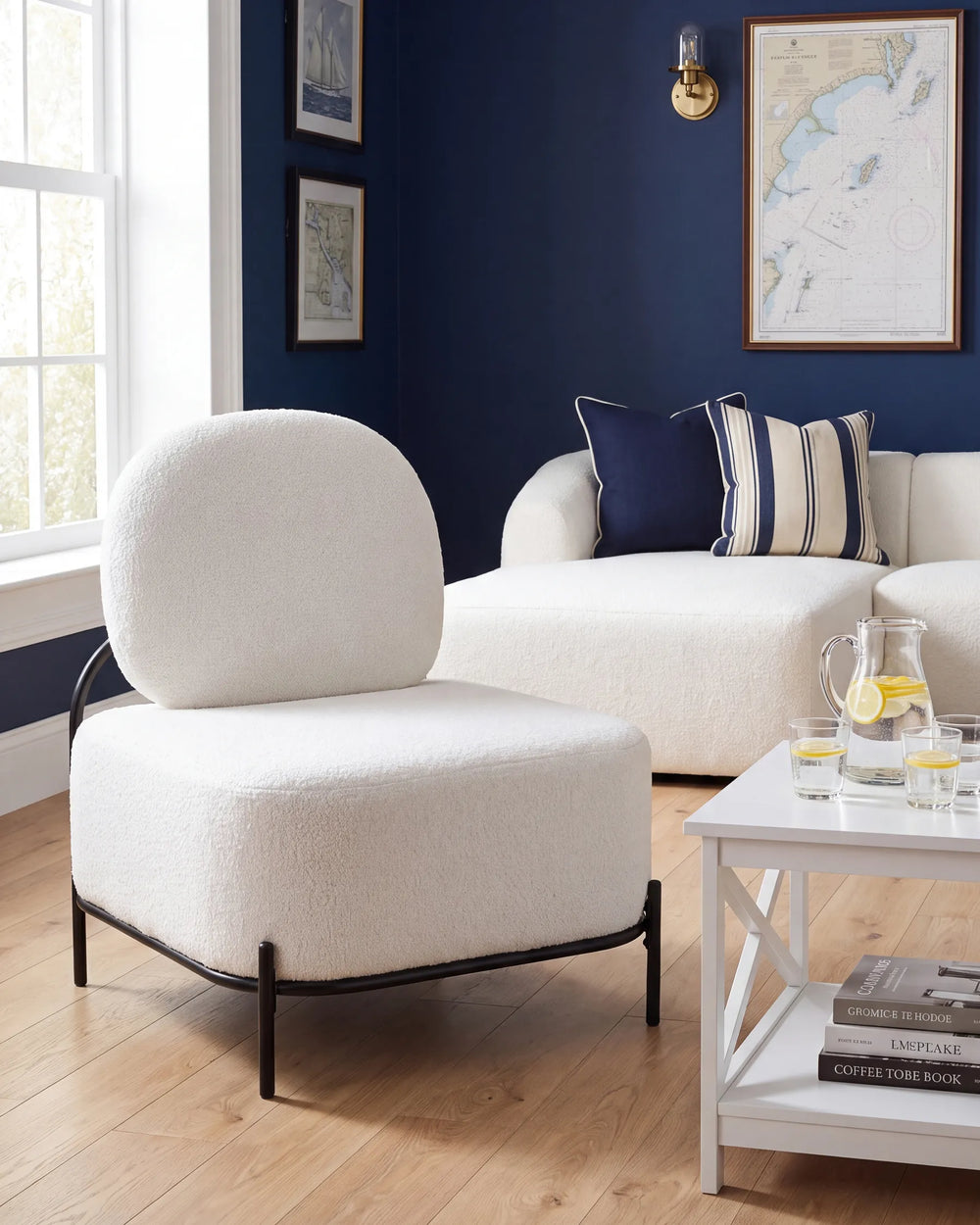 A modern white bouclé lounge chair with a black metal frame, positioned in a living room setting with a navy blue wall, a white sofa, and a small white side table.